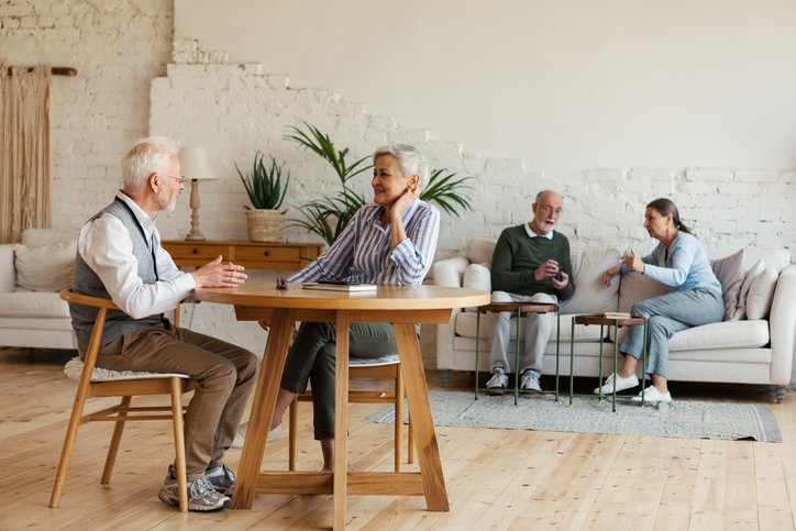 Senior man and woman sitting at table and enjoying talk, another aged couple interacting in background sitting on sofa in common room of nursing home