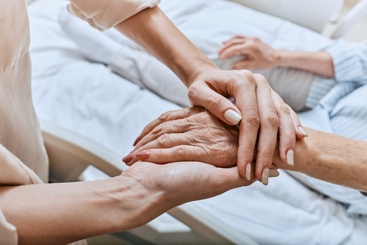 Relative holding trembling hand of senior woman with Parkinson's disease lying in hospital bed at medical ward. Diagnosis and treatment of Parkinson's disease and dementia